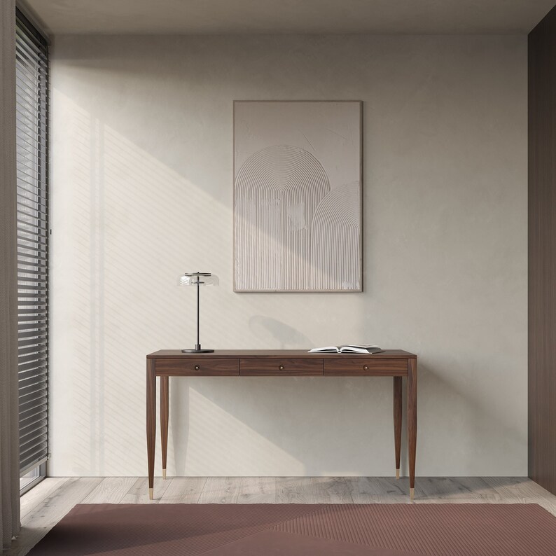 May include: A dark wood console table with three drawers and brass legs. The table is in a room with a white wall and a framed abstract print above it.
