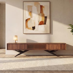 May include: A modern living room featuring a wooden media console with a lamp, a large abstract painting, and a potted plant. The console has a slatted center section and sits on black metal legs. A cream-colored rug covers the wooden floor.