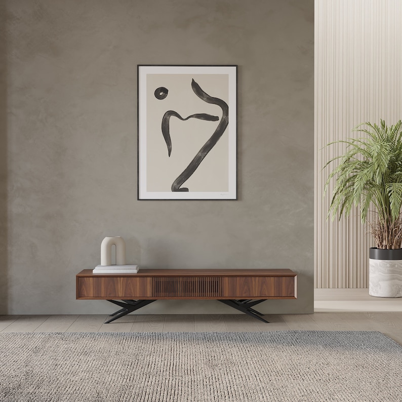 May include: A contemporary living room with a dark wood media unit and black metal legs. Above, a framed abstract art print with black ink lines on a beige background. A potted plant and a rug complete the decor, creating a stylish space.