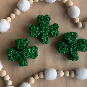 May include: Three green shamrock-shaped treats made of puffed rice cereal, on a brown background with a white and brown beaded garland.