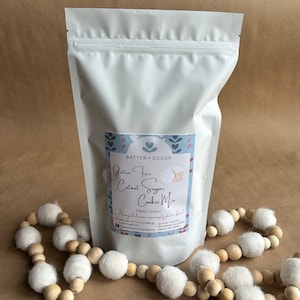 May include: A white resealable bag with a label that says "Gluten-Free Cutout Sugar Cookie Mix" and "Makes 2 Dozen". The label has a blue and white floral design and a picture of sugar cookies.