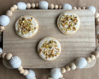 May include: Three round sugar cookies with white icing and chopped pistachios on top. The cookies are arranged on a wooden cutting board with a white and brown beaded garland around the edge.