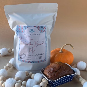 May include: A white bag of gluten-free pumpkin bread mix with a blue and pink label that says "Gluten-Free Pumpkin Bread Mix" and "Always Delicious, amazingly gluten free!"  A loaf of pumpkin bread in a blue and white patterned tin, a small orange pumpkin, and a garland of wooden beads with white pom-poms.