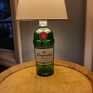 May include: A unique table lamp crafted from a green Tanqueray gin bottle, topped with a beige lampshade. The lamp sits on a round, wooden table, creating a distinctive home decor piece.