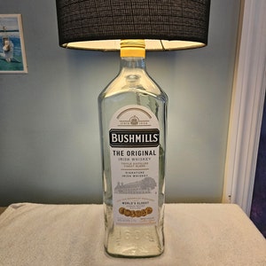 May include: A clear glass bottle lamp with a black lampshade. The bottle is labelled "Bushmills The Original Irish Whiskey" and "World's Oldest Licensed Whiskey Distillery".