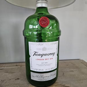 May include: A green glass lamp base made from a Tanqueray London Dry Gin bottle. The bottle has a white label with black text and a red wax seal. The lamp has a white lampshade.