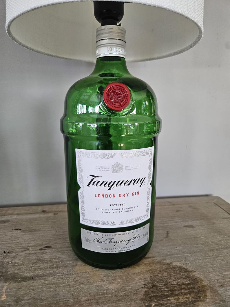 May include: A green glass lamp base made from a Tanqueray London Dry Gin bottle. The bottle has a white label with black text and a red wax seal. The lamp has a white lampshade.