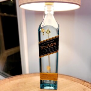 May include: A clear glass bottle lamp with a gold label that reads "Johnnie Walker Blue Label Blended Scotch Whisky A Blend of Our Rarest Whiskies" and a beige lampshade. The bottle is sitting on a wooden surface.