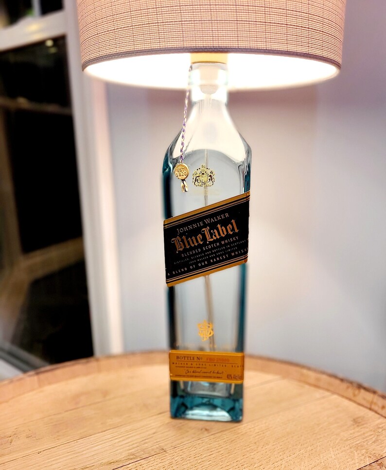 May include: A clear glass bottle lamp with a gold label that reads "Johnnie Walker Blue Label Blended Scotch Whisky A Blend of Our Rarest Whiskies" and a beige lampshade. The bottle is sitting on a wooden surface.