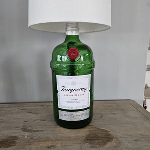 May include: A green glass lamp base made from a Tanqueray London Dry Gin bottle. The bottle has a white label with black text and a red wax seal. A white lampshade sits atop the bottle.