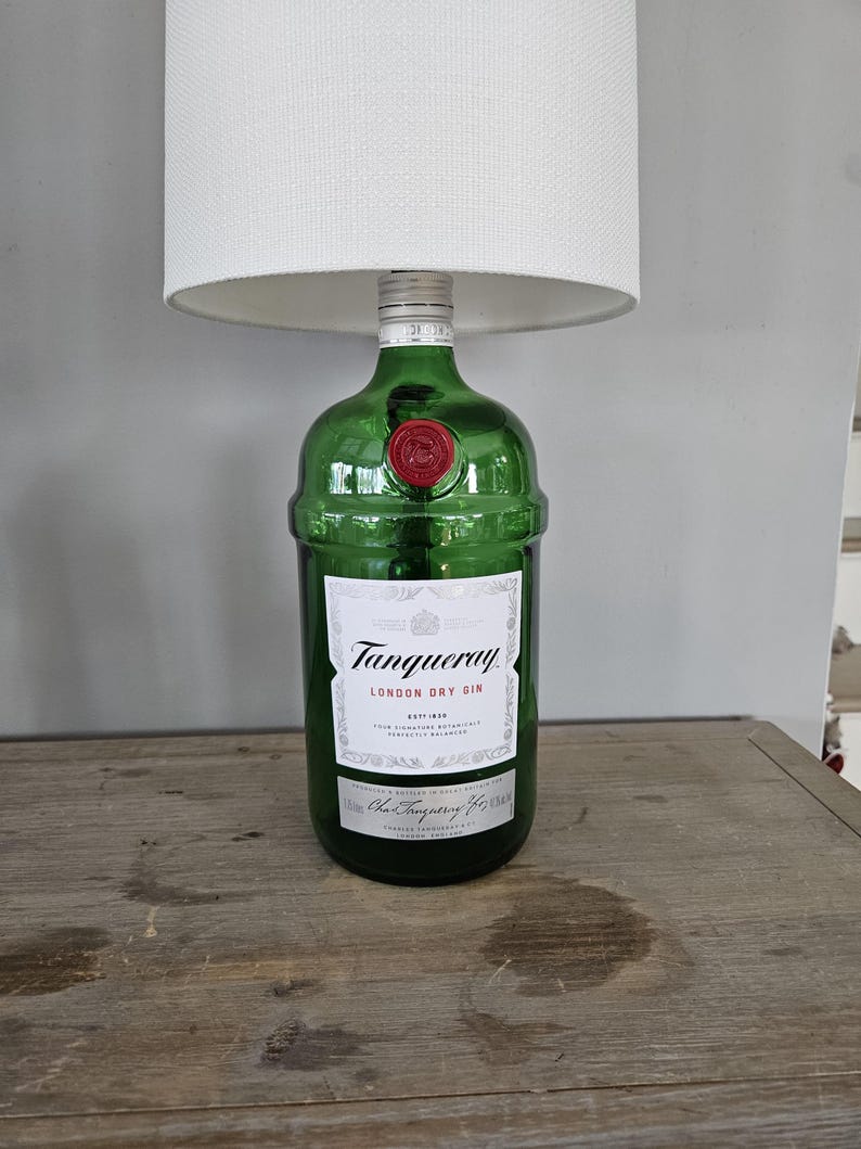 May include: A green glass lamp base made from a Tanqueray London Dry Gin bottle. The bottle has a white label with black text and a red wax seal. A white lampshade sits atop the bottle.