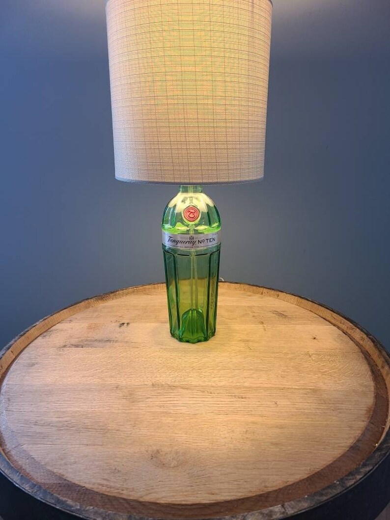 May include: A custom-made lamp featuring a green glass bottle base with the text "Tanqueray No. TEN". The lamp has a beige lampshade and is placed on a wooden barrel. The lamp is lit, casting a warm glow.