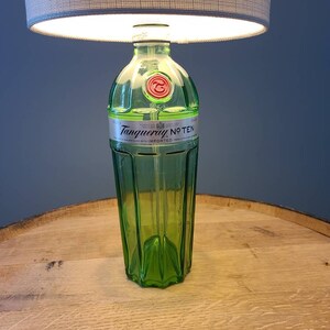 May include: A repurposed lamp made from a green glass gin bottle, featuring the "Tanqueray No. TEN" label. The lamp has a gray lampshade and sits on a wooden surface. The lamp is a unique home decor item.