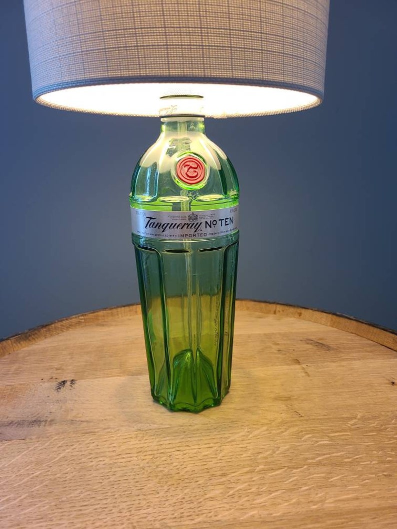 May include: A repurposed lamp made from a green glass gin bottle, featuring the "Tanqueray No. TEN" label. The lamp has a gray lampshade and sits on a wooden surface. The lamp is a unique home decor item.