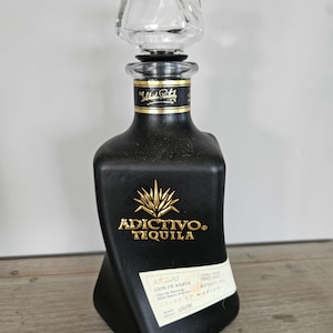 May include: A matte black tequila bottle with a twisted square base and a clear, sculpted glass stopper. The bottle features gold embossed text reading ADICTIVO TEQUILA and an agave plant icon. A white label displays 750 ml and 40% alc. vol.
