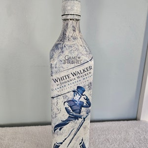 May include: A white glass bottle with a blue label that says "Game of Thrones White Walker by Johnnie Walker Blended Scotch Whisky Limited Edition". The bottle has a white and blue design with a figure of a man in a blue coat and black hat holding a staff.
