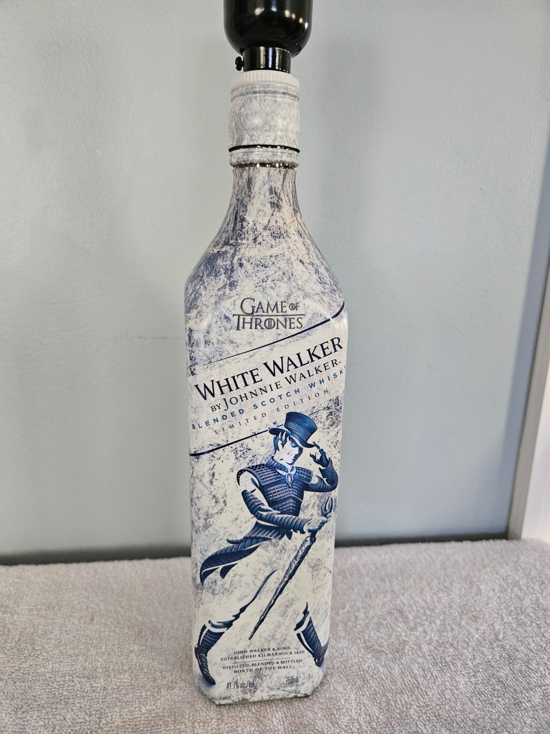 May include: A white glass bottle with a blue label that says "Game of Thrones White Walker by Johnnie Walker Blended Scotch Whisky Limited Edition". The bottle has a white and blue design with a figure of a man in a blue coat and black hat holding a staff.