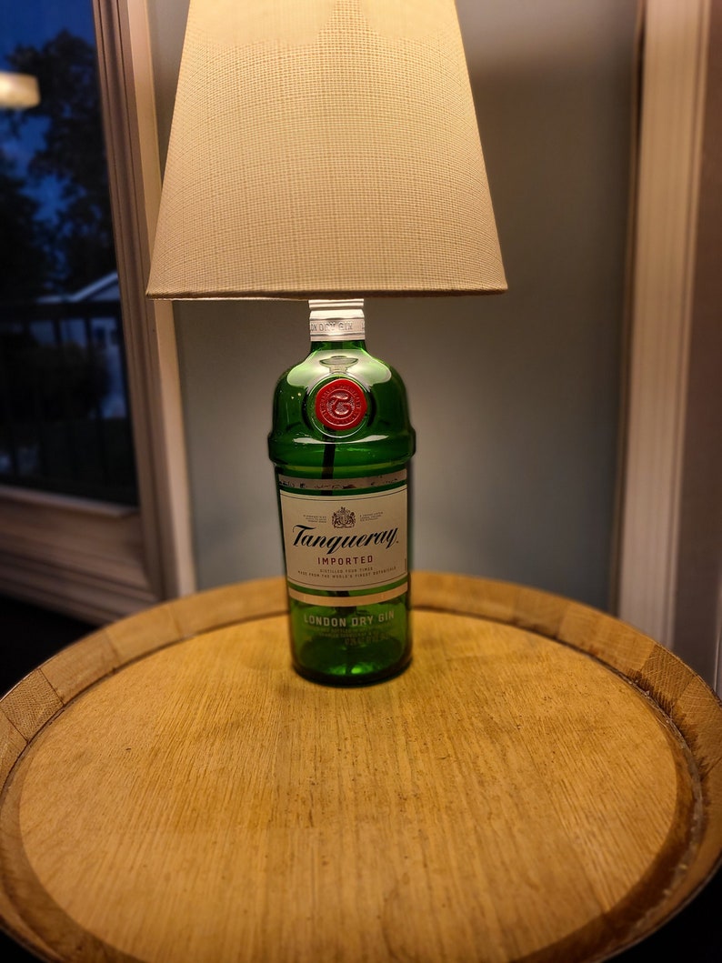 May include: A unique table lamp crafted from a green Tanqueray gin bottle, topped with a beige lampshade. The lamp sits on a round, wooden table, creating a distinctive home decor piece.