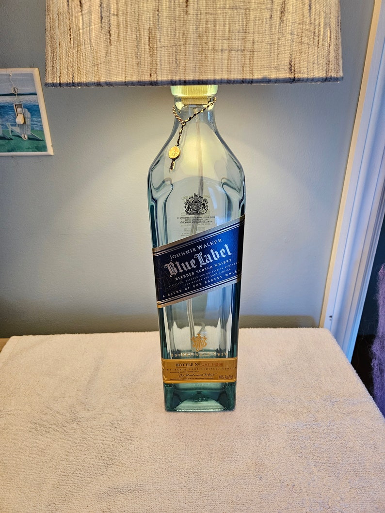 May include: A clear glass bottle lamp with a blue and gold label that reads "Johnnie Walker Blue Label Blended Scotch Whisky". The bottle is sitting on a white surface.