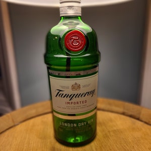 May include: A green Tanqueray London Dry Gin bottle with a red wax seal. The bottle is on a wooden surface. The label reads "IMPORTED" and "LONDON DRY GIN". The bottle is under a lamp.