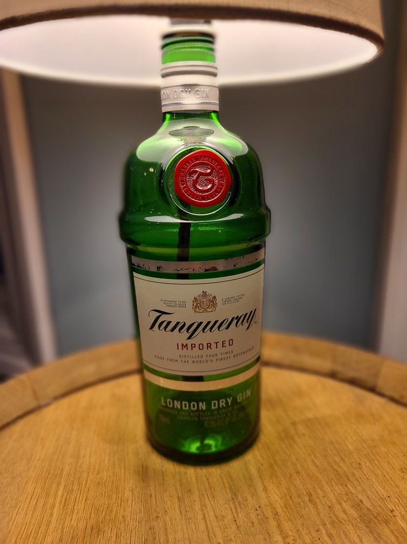 May include: A green Tanqueray London Dry Gin bottle with a red wax seal. The bottle is on a wooden surface. The label reads "IMPORTED" and "LONDON DRY GIN". The bottle is under a lamp.