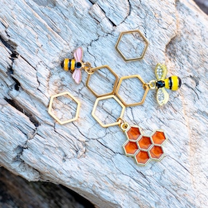 Könnte beinhalten: Goldfarbene Bienenanhänger mit einem Honigwabenanhänger, alle an goldfarbenen sechseckigen Ringen hängend. Die Anhänger befinden sich auf einer hellbraunen Holzoberfläche.