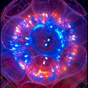 May include: A clear plastic sphere with multiple cup-shaped compartments, each containing a small LED light. The lights are arranged in a circular pattern and emit a mix of blue and orange light.