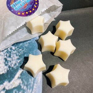 May include: A white paper bag with a purple and yellow star-shaped sticker that says "Star Melt Creations" is open and spilling out six white star-shaped wax melts.