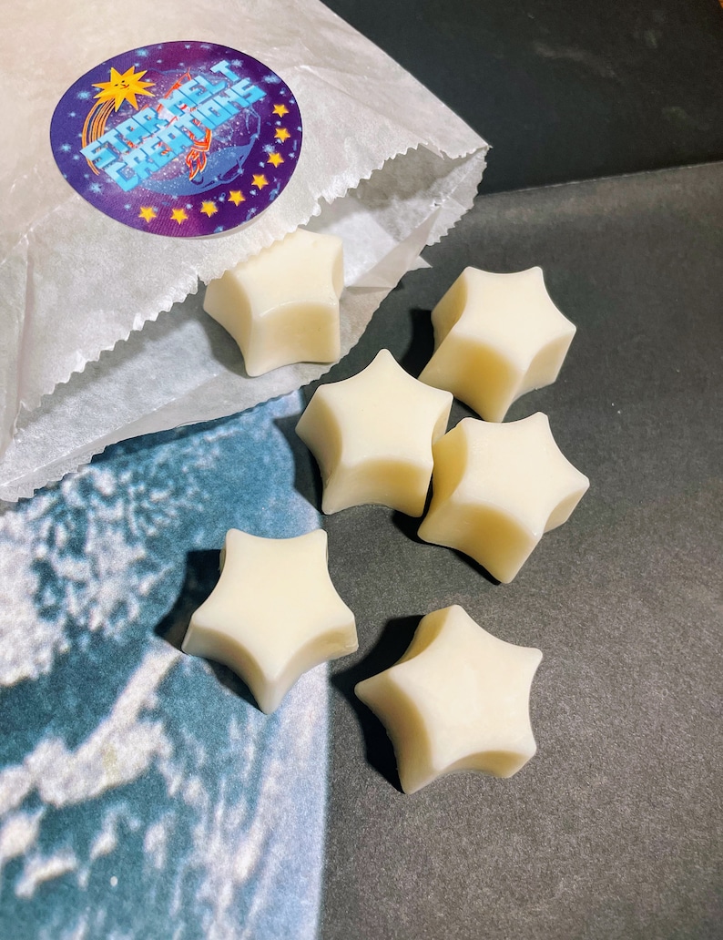 May include: A white paper bag with a purple and yellow star-shaped sticker that says "Star Melt Creations" is open and spilling out six white star-shaped wax melts.