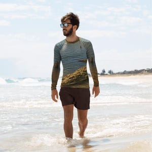 May include: A long-sleeve rash guard shirt featuring a photographic print of a coastal landscape with blue ocean water and a sandy shoreline. The shirt has a crew neckline and raglan sleeves, paired with solid dark brown swim trunks.