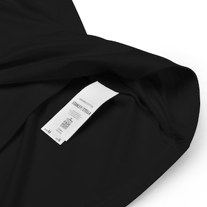 Op de afbeelding: Een wit label bevestigd aan een zwart T-shirt. Het label vermeldt "ORGANIC COTTON STANLEY/STELLA" en bevat een Fair Wear Foundation-logo. Het label geeft ook de maat "M" en de borstmaat van 97/99 cm aan.