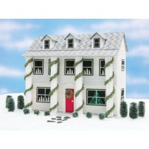 May include: A white dollhouse with a red door and green garland decorations around the columns. The house has a gray roof and windows with lit candles inside. There are small green trees in front of the house.