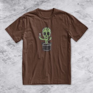May include: A brown t-shirt with a green cactus with a skull face and a bone in its hand. The cactus is in a black pot.