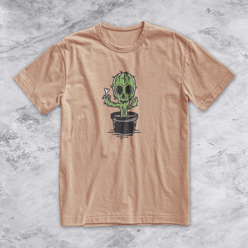 May include: A tan t-shirt with a green cactus wearing a skull and holding a bone. The cactus is in a black pot.