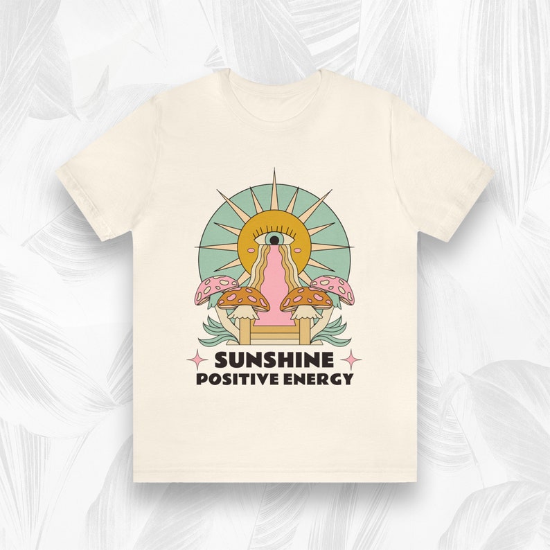 Peut inclure: Un t-shirt beige avec un motif graphique repr&eacute;sentant un soleil avec un &oelig;il, une cascade et des champignons. Le texte "SUNSHINE POSITIVE ENERGY" est imprim&eacute; sous le motif.