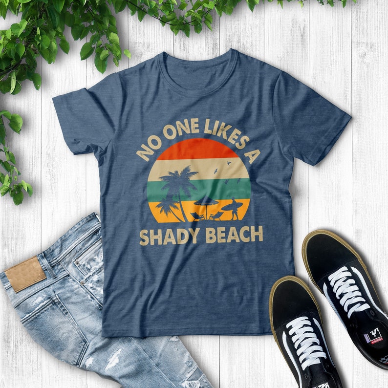 May include: A navy blue t-shirt with a vintage-style graphic of a palm tree and a surfer on a beach. The graphic is surrounded by a circle with the text "NO ONE LIKES A SHADY BEACH".