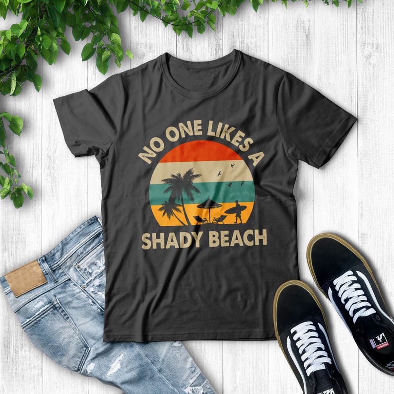 May include: A black t-shirt with a vintage-style graphic of a palm tree and a surfer on a beach. The graphic is surrounded by a circle with the text "No one likes a shady beach".
