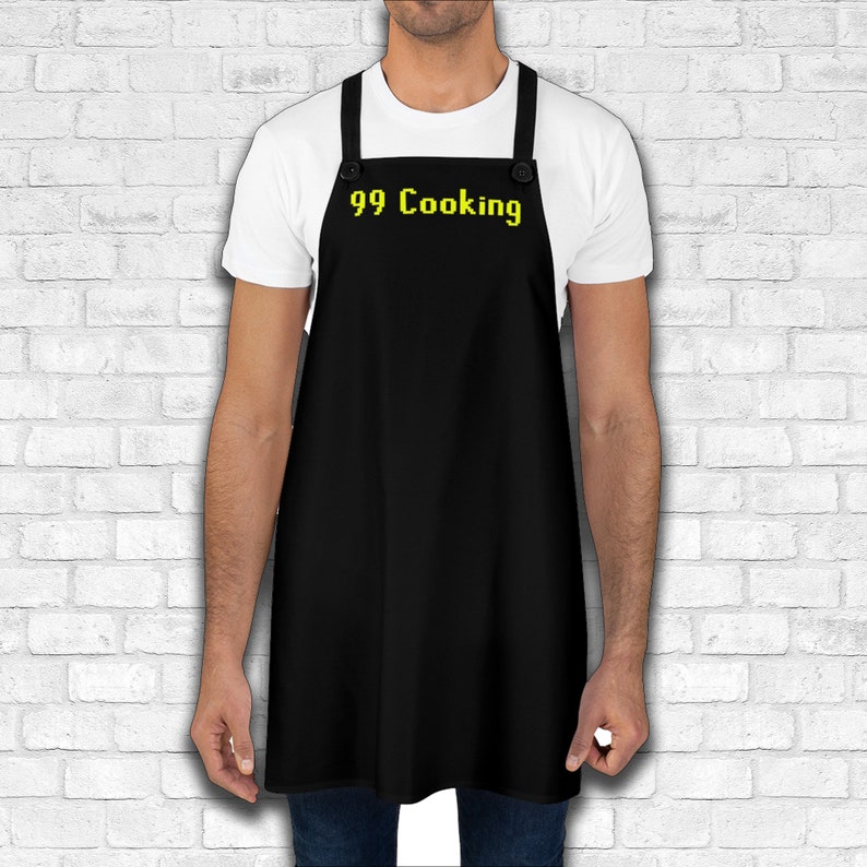 K&ouml;nnte beinhalten: Eine schwarze Sch&uuml;rze mit verstellbaren Tr&auml;gern und zwei Kn&ouml;pfen. Die Sch&uuml;rze tr&auml;gt den Text "99 Cooking" in Gelb.
