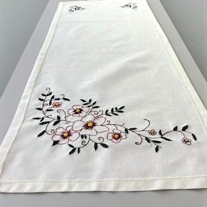 May include: Cream-colored embroidered table runner with a floral design. The embroidery features burgundy, yellow, and green flowers and leaves. The runner is rectangular and appears to be made of linen or a similar fabric.