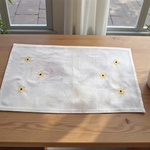 May include: A rectangular white table runner with embroidered yellow and black flower designs. The runner is placed on a wooden table, with a potted plant on the side. The runner is approximately 18 inches wide and 30 inches long.