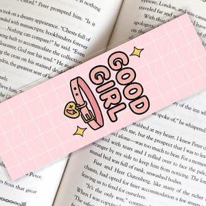 Pode incluir: Marcador de livros rosa com as palavras "GOOD GIRL" em letras pretas e rosa. O marcador apresenta uma coleira rosa com um pingente em forma de cora&ccedil;&atilde;o e detalhes dourados. Duas estrelas douradas tamb&eacute;m est&atilde;o inclu&iacute;das.