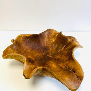 May include: A large, organically shaped wooden bowl with a natural, flowing design. The bowl is crafted from a light brown wood, showcasing intricate grain patterns and a smooth, polished finish. The edges undulate, creating a unique, sculptural form.