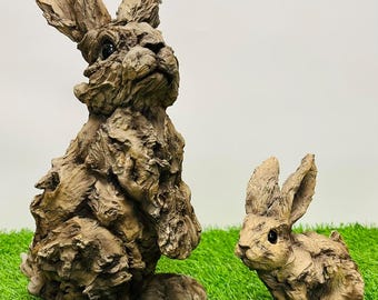 May include: Two brown, wooden rabbit sculptures on a green artificial grass surface. The larger rabbit is standing upright with its front paws raised. The smaller rabbit is sitting with its head tilted to the side.