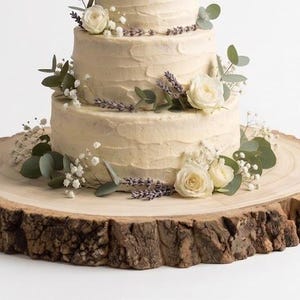 May include: A three-tiered cake with off-white textured frosting, decorated with sprigs of lavender, white roses, and eucalyptus leaves. The cake rests on a circular wooden slab featuring a natural bark edge and visible wood grain rings.