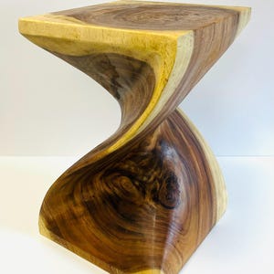 May include: A handcrafted wooden stool with a unique, twisted hourglass design. The stool features a square top and base, with a smooth, curved body. The wood grain is visible, showcasing natural brown and tan tones. The stool is a decorative accent piece.