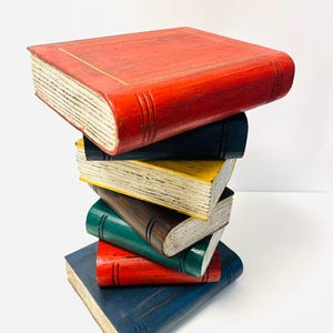 May include: A stack of six decorative wooden books in various colours: red, navy blue, yellow, brown, green, and red. The books have aged, off-white pages and are stacked on top of each other.