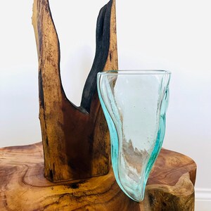 May include: A clear, light blue glass vase with a unique, organic shape. The vase sits on a rustic wooden table next to a tall, dark brown wooden sculpture. The sculpture has a natural, split design with a charred black section.