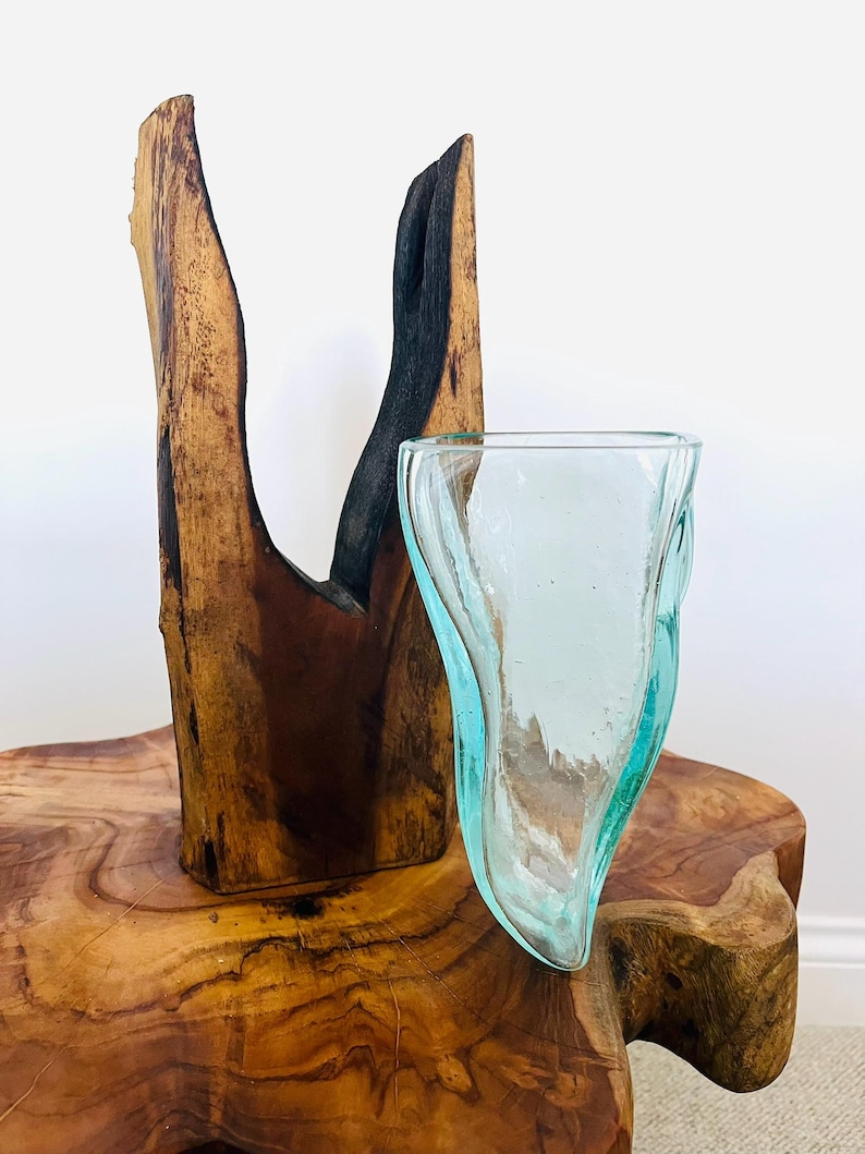 May include: A clear, light blue glass vase with a unique, organic shape. The vase sits on a rustic wooden table next to a tall, dark brown wooden sculpture. The sculpture has a natural, split design with a charred black section.