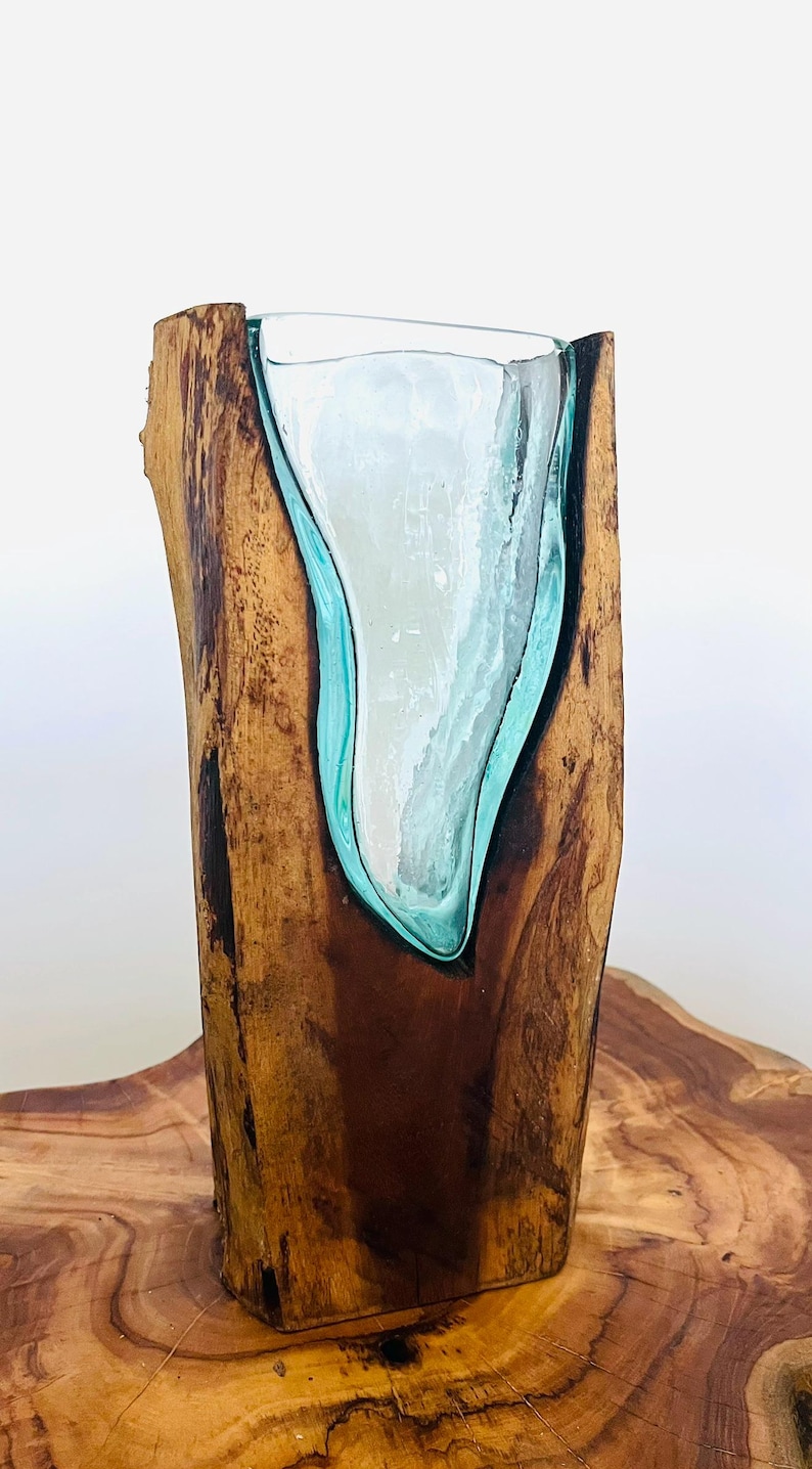 May include: A handcrafted wooden vase with a clear glass insert. The vase is made from a single piece of wood, with a natural, organic shape. The glass insert has a teal-colored border and a white interior, creating a striking contrast. The vase is displayed on a wooden surface.