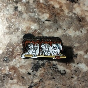 May include: A white enamel pin with a cartoon image of a s'more with a chocolate bar, graham crackers, and marshmallows. The s'more is melting and dripping chocolate.